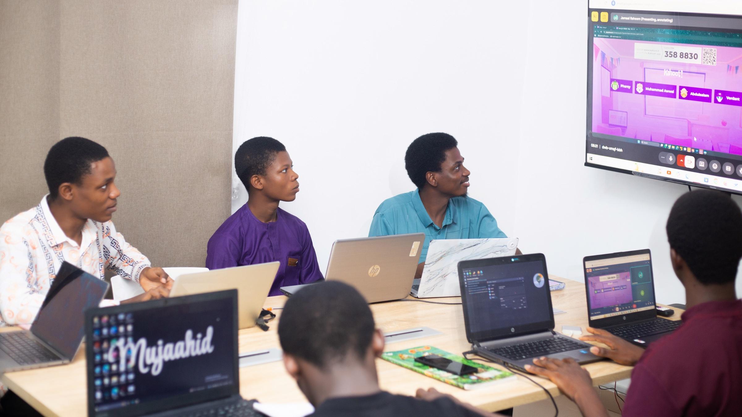 Students at a shared table with laptops, looking up at a quiz on the classroom screen
